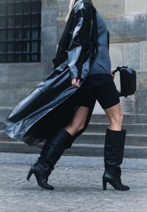 Un manteau en cuir noir drape sur un pull gris, associé à un short de vélo noir et des bottes noires jusqu'au genou, mettant en avant un style élégant et moderne.