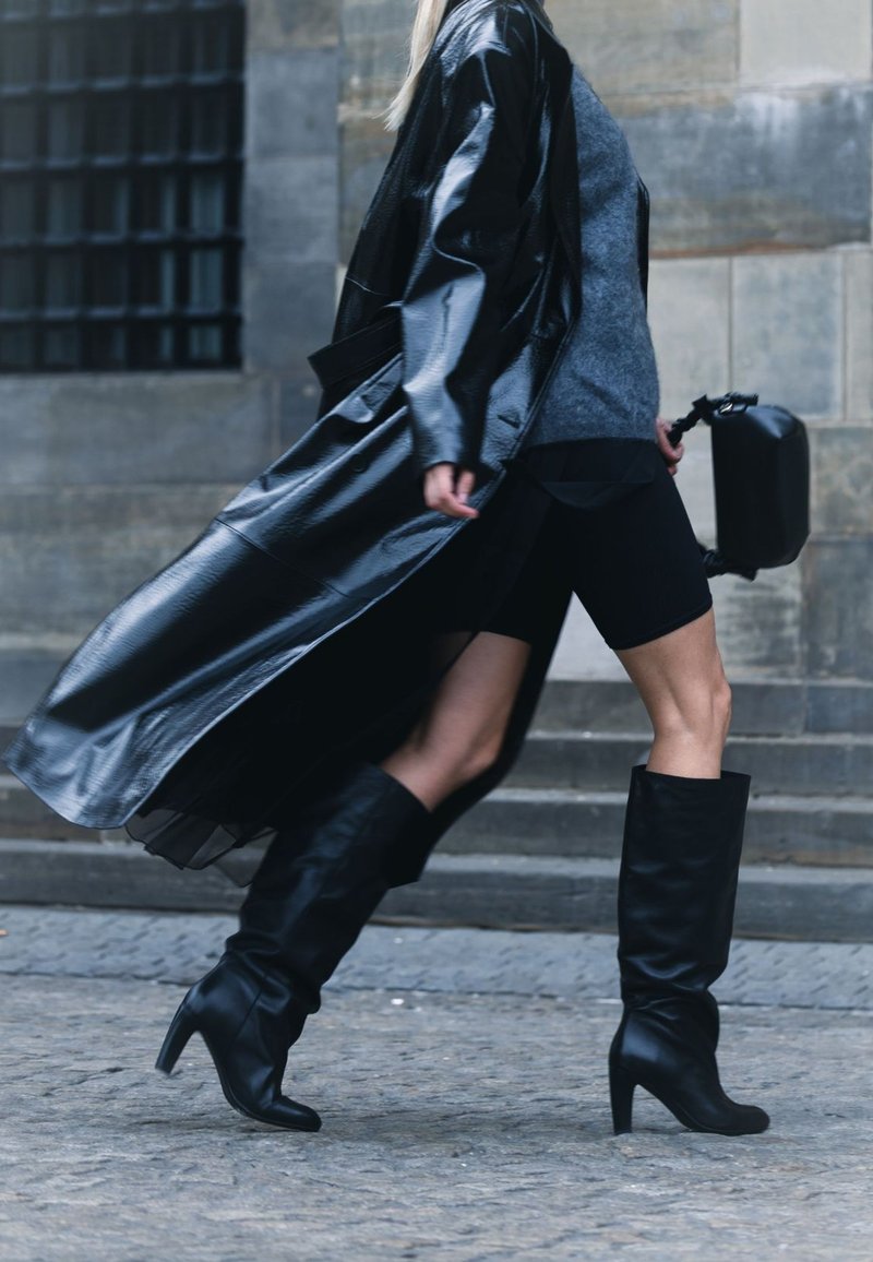 Un manteau en cuir noir drape sur un pull gris, associé à un short de vélo noir et des bottes noires jusqu'au genou, mettant en avant un style élégant et moderne.
