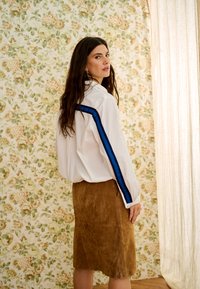 Vrouw met lang donker haar, een wit shirt met blauwe strepen op de mouwen en een bruine suède rok, staand bij bloemenbehang en een doorschijnend gordijn.