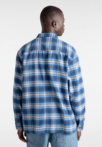 Mies, który ma na sobie niebiesko-biały w kratę koszulę z długim rękawem oraz jasnoniebieskie dżinsy, pozujący tyłem do aparatu na białym tle.