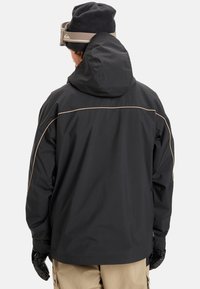 Juodas vandeniui atsparus paltas su kaukole, su smėlio spalvos apdaila siūlėse ir laisvu siluetu. Dėvimi su smėlio spalvos kelnėmis ir pirštinėmis.