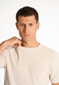 Camisa corta de manga con textura, de color beige claro y escote redondo. La persona está ajustando la tela cerca del hombro. Fondo limpio.