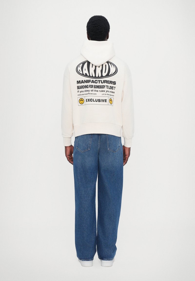 Krämfärgad hoodie med svart grafisk text på baksidan, i kombination med blå vida jeans. Vita sneakers fulländar outfiten.