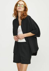 Camisa negra de botones con mangas cortas y un tejido texturizado, combinada con pantalones cortos negros. El modelo lleva gafas de sol y una camiseta sin mangas blanca.