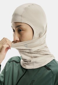 Person adjusting beige balaclava-style headwear over green jacket, focused on pulling fabric over lower face.