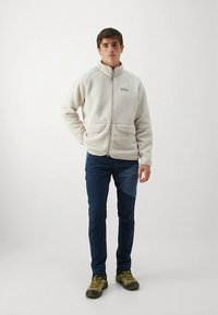 Jasno beżowa kurtka z polaru z podwyższonym kołnierzem, przednimi kieszeniami i zamkiem błyskawicznym. Noszona z granatowymi spodniami i brązowymi butami trekkingowymi. Prosty design, casualowy krój.