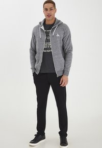 Sivi zip-up dukserica s bijelim logotipom, u kombinaciji s tamnosivim majicom s printom i crnim uskim hlačama. Crne tenisice upotpunjuju izgled.