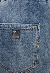Jean en denim avec une poche arrière portant une étiquette noire. Couleur : bleu moyen avec des surpiqûres orange. Texture : lisse avec une légère brillance.