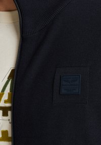 Temno modra zip-up jopica z ribastim ovratnikom in teksturo blaga, z kvadratnim logotipom na levi strani. Prelita čez bež majico.
