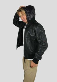 Homme blond portant une veste en cuir noir avec capuche, un pantalon beige et une chemise blanche, posant avec une main sur la tête devant un fond uni.