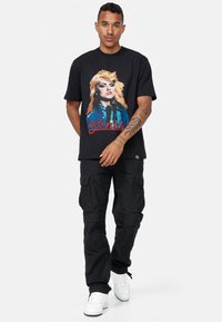 Černé bavlněné tričko s barevným grafickým motivem osoby se světlými vlasy a textem "Blondie". V kombinaci s černými cargo kalhotami a bílými teniskami.