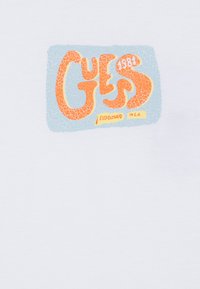 Biele tričko s textúrovanou grafiku v oranžovej a modrej farbe, s štýlovým textom "GUES 1984" a "ESTABLISHED IN L.A." v žltej farbe.