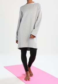 Vestido suéter gris con mangas largas, escote redondeado y abertura lateral. Lleva puesto un legging negro sobre una esterilla de yoga rosa.