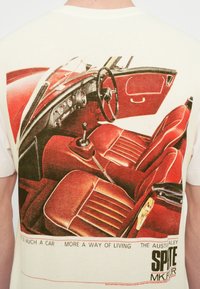 Βαμβακερό μπλουζάκι με σχέδιο που απεικονίζει το εσωτερικό ενός κόκκινου αυτοκινήτου, παρουσιάζοντας λεπτομερέστατα τα καθίσματα, τον πίνακα οργάνων και το λεβιέ ταχυτήτων, με κείμενο κάτω από την εικόνα.