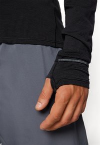 Haut à manches longues noir avec texture côtelée et poignets avec trou pour le pouce, associé à un short de sport gris avec une finition en tissu lisse.