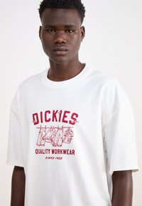 T-shirt di cotone bianco con una stampa grafica rossa di articoli di abbigliamento da lavoro appesi, con il testo "DICKIES" e "QUALITY WORKWEAR". Maniche corte.