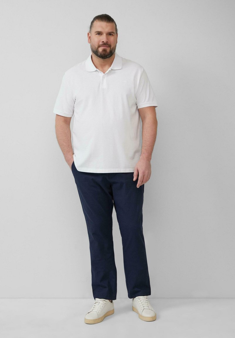 Mann mit Bart, der ein weißes Polohemd, dunkelblaue Hose und weiße Sneakers trägt, steht vor einer schlichten grauen Wand mit einer Hand in der Tasche.