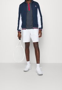 Marinblå zip-up jacka med vita sidopaneler, randig polotröja, vita shorts och vita sneakers, håller en tennisracket.