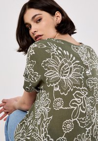 Blusa verde olivo con estampado floral blanco, escote redondo modesto, mangas cortas y detalles de botones en la parte posterior.