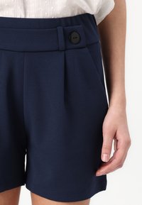 Námořnické modré šortky vyrobené z hladké látky, s plisovaným designem, boční kapsou a černým knoflíkem jako akcentem na pase.