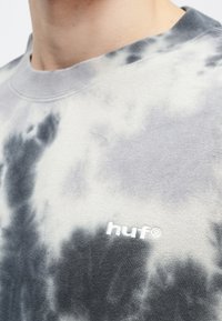 Sweatshirt cinzento com efeito tie-dye, apresentando um colarinho canelado e um discreto bordado branco "huf" no peito. Textura suave com padrões de cor variados.