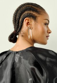 Sidabriniai auskarai su kaspinėlio dizainu ir ilgais, tiesiais akcentais. Viršus yra juodas satinas, turintis lygų paviršių ir subtilius raštus.