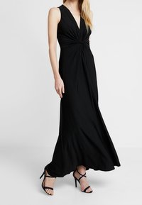 Musta, hihaton maksimekko, jossa on syvä V-kaula-aukko ja solmuvyötärö, valmistettu sileästä kankaasta, yhdistetty mustien, hihnallisten korkokenkien kanssa.