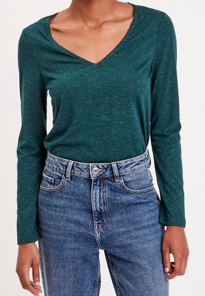Personne portant un haut vert foncé à manches longues avec un col en V, rentré dans un jean en denim bleu taille haute avec des poches avant visibles et un bouton.