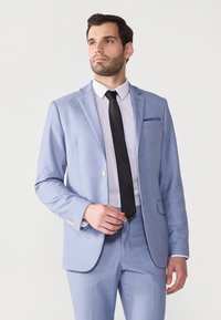 Heller blauer Anzug aus strukturiertem Stoff, mit einer Ein-Knopf-Verschluss, Reverskragen und einer Brusttasche. Kombiniert mit einer schwarzen Krawatte.
