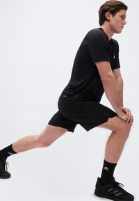 Homme portant une tenue de sport noire, étirant la jambe gauche vers l'avant, tenant le genou droit, debout devant un fond blanc uni.