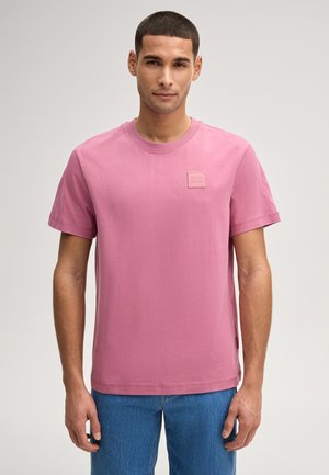 Mann trägt ein rosa kurzärmeliges T-Shirt mit einem kleinen "JOOP! JEANS"-Aufnäher auf der Brust und blaue Jeans, steht vor einem einfarbigen Hintergrund.