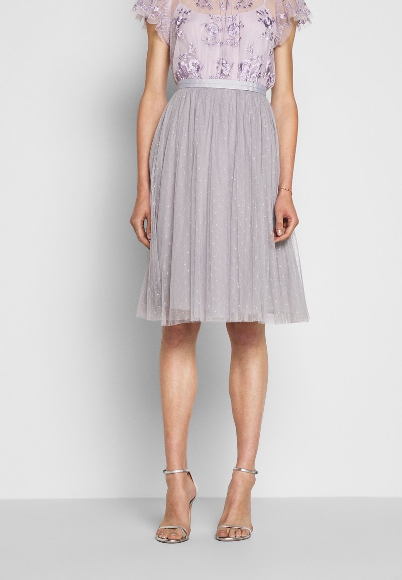 Femme portant un chemisier floral violet clair rentré dans une jupe midi gris clair transparent à pois, avec des talons argentés à bride à la cheville.