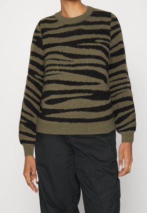 Personne portant un pull zébré kaki et noir et un pantalon cargo noir, debout devant un fond clair uni.