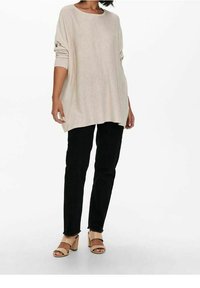 Femme portant un haut beige ample à manches longues, un pantalon noir droit et des sandales à talons beiges, debout devant un fond blanc.