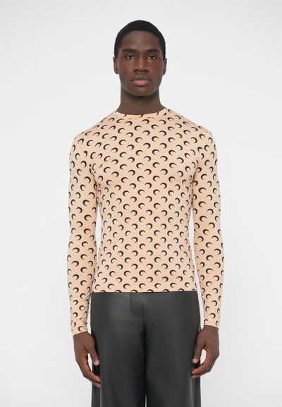 Haut à manches longues couleur pêche clair avec motif de croissant de lune noir ; coupe ajustée, texture lisse et col rond. Associé à un pantalon noir.