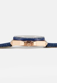 Blå denimrem klocka med ett roseguld åttakantigt boett, en knapp på sidan och en texturerad bezel. Glasansikte med en ren urtavla.