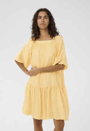 Kobieta o kręconych, ciemnych włosach, ubrana w luźną, krótkorękawową, jasnofioletową sukienkę z marszczonym pasem, stojąca na tle gładkiego szarego tła.