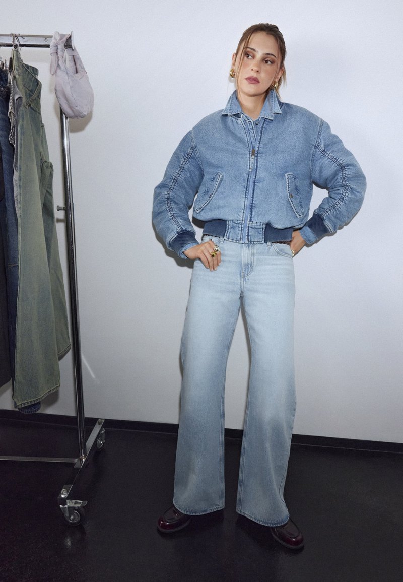 Frau steht mit den Händen in den Hüften, trägt eine blaue Jeansjacke, weit geschnittene hellblaue Jeans und dunkle Schuhe neben einem Kleiderständer.