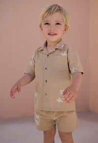 Camicia corta color marrone chiaro con ricamo di palme e pantaloni abbinati, realizzati in un tessuto morbido. Presenta una tasca frontale e bottoni.