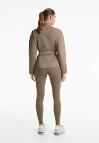 Kanca lófarokkal, taupe színű, hosszú ujjú, övvel ellátott dzsekit, hozzáillő leggingst és fehér sportcipőt visel, háttal állva fehér háttér előtt.