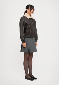 Woman wearing a dark brown leather jacket with a cream shearling collar, grey pleated skirt, black sheer tights, and black flats, standing sideways.