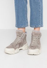 Harmaat mokkanahka nilkkurit, joissa on teksturoitu pinta, valkoinen kumipohja ja tekoturkiskaulus, etulenkki ja sivuvetoketju.