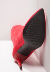 Røde fløjlsstiletto støvler med spids tå og en tekstureret sort gummisål. Designet med en tætsiddende silhuet, der går over læggen.