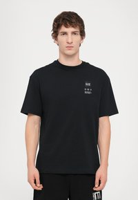 Melnā kokvilnas T-krekls ar īsām piedurknēm, apaļu kakla izgriezumu un nelielu drukātu etiķeti kreisajā krūšu pusē. Gluda tekstūra, brīvs piegriezums.