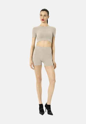 Frau mit zurückgebundenem Haar, trägt beige kurzärmliges Crop-Top und passende hoch taillierte Shorts, schwarze spitze High Heels, stehend vor weißem Hintergrund.
