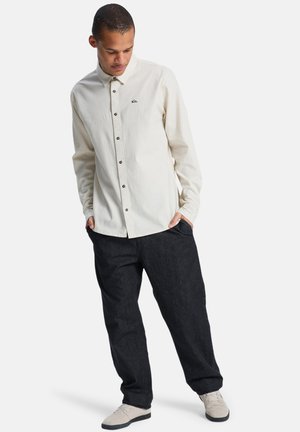 Lys beige skjorte med knapper og krave samt et logo på brystet, parret med mørke denimbukser og lyse sko.