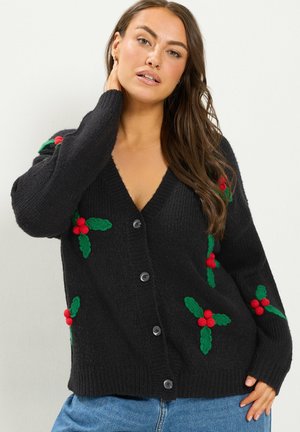 Femme portant un cardigan noir avec une broderie de houx rouge et verte, un jean bleu à jambes larges et des ballerines noires, debout devant un fond uni.
