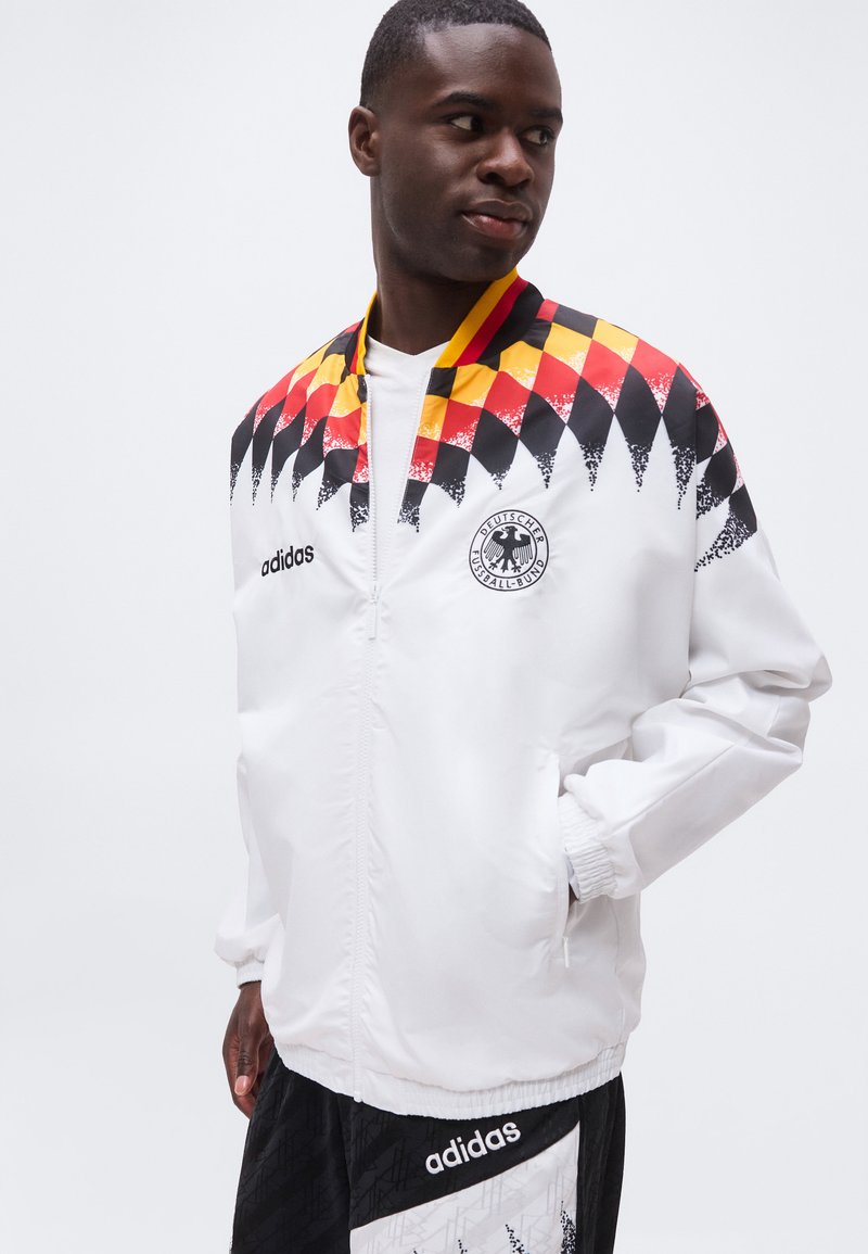 Mann mit weißer Adidas-Jacke mit rot-gelb-schwarzem Schachbrettmuster und Deutscher Fussball-Bund-Logo, Hände in den Taschen, schaut seitlich.