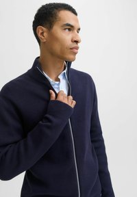 Tamsiai mėlynas užtrauktuku užsegamas švarkas pagamintas iš tekstūruotos medžiagos, turi aukštą apykaklę ir gumbuotas rankogalius. Užtrauktukas turi kontrastingą sidabrinę apdailą.