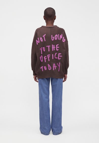 Personne portant un pull marron avec un texte rose "Not going to the office today" dans le dos et un jean bleu ample, debout, dos à nous.